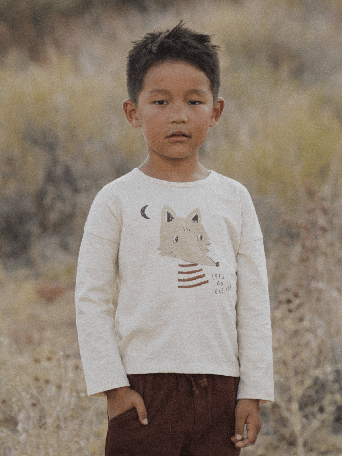 Child wearing a light shirt with a fox design, standing outdoors amidst tall grass and soft, neutral tones.