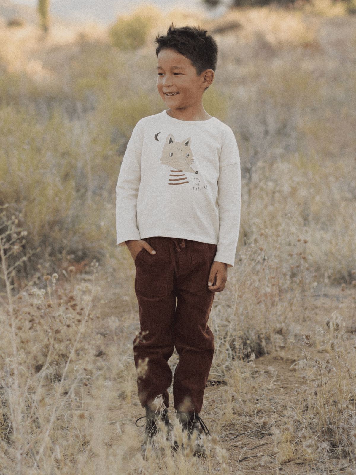 Smiling boy in a nature setting, wearing a t-shirt with a fox design and brown pants, surrounded by tall grass.