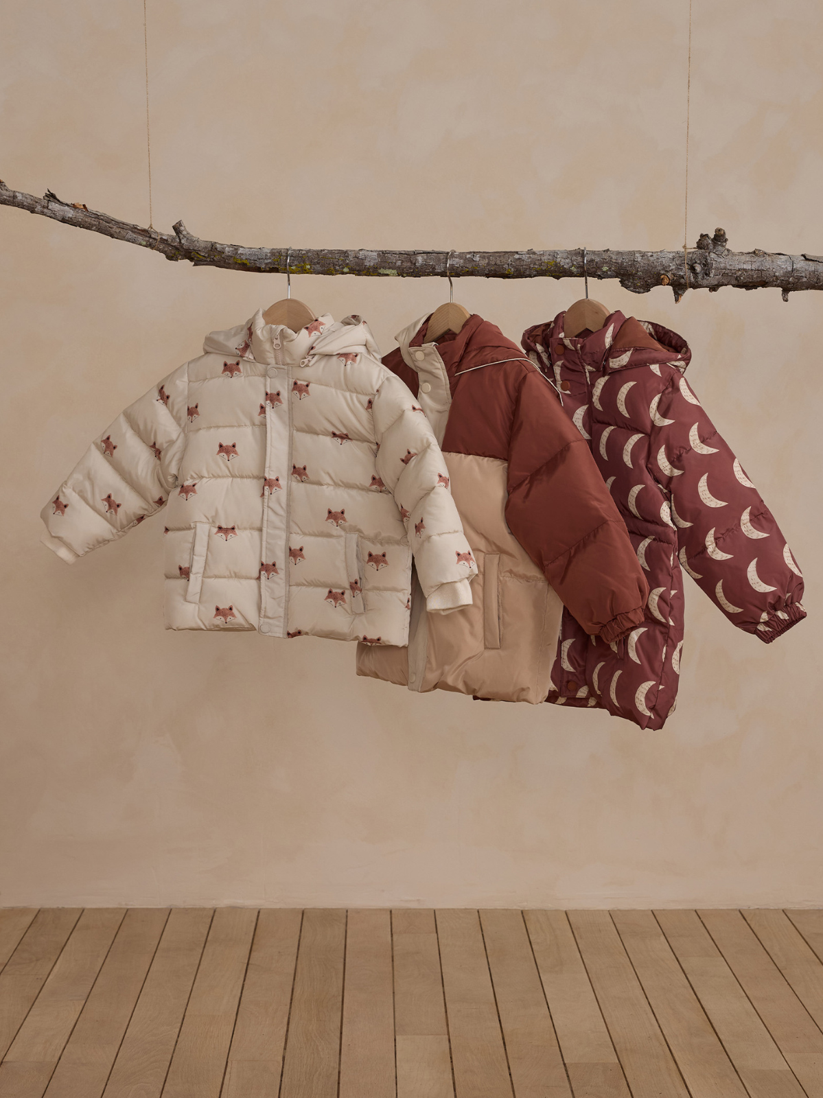 Three puffer jackets in various colors and patterns hanging from a wooden branch against a light background.