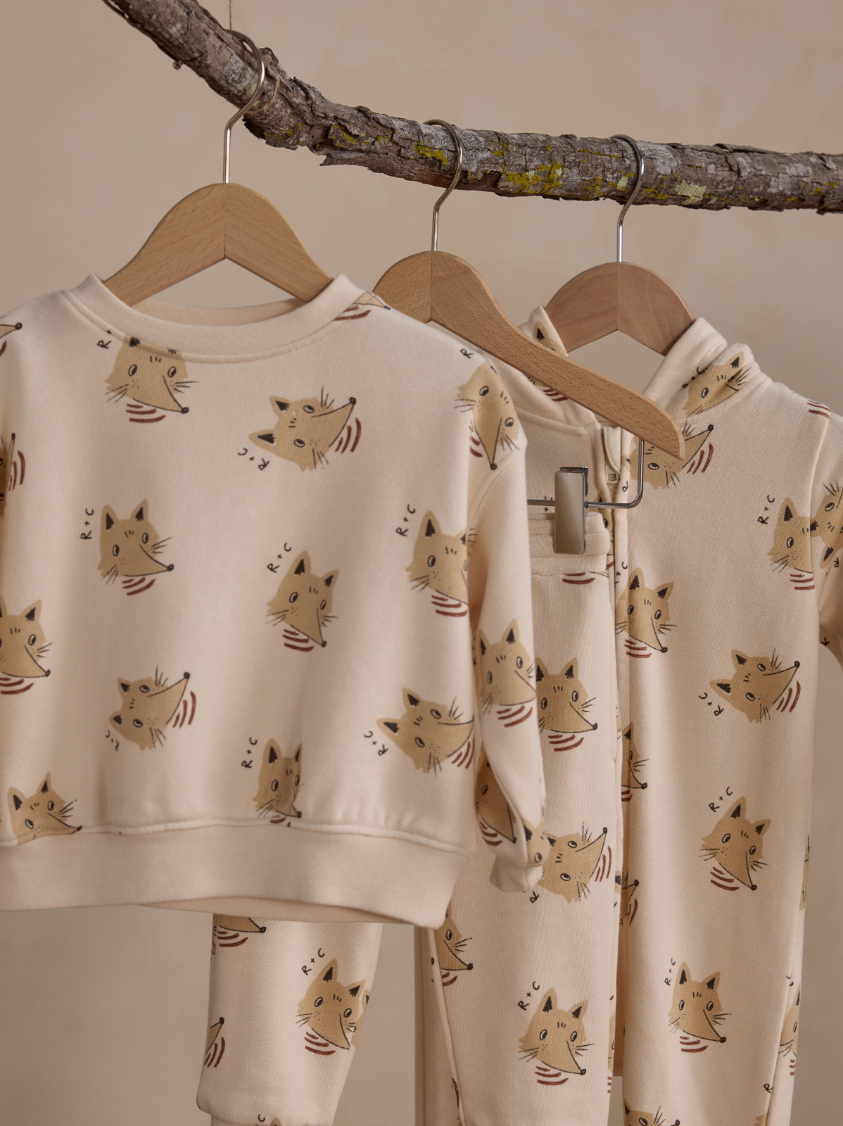 Children's sweatshirts with playful fox patterns, hanging on wooden hangers against a soft beige background.