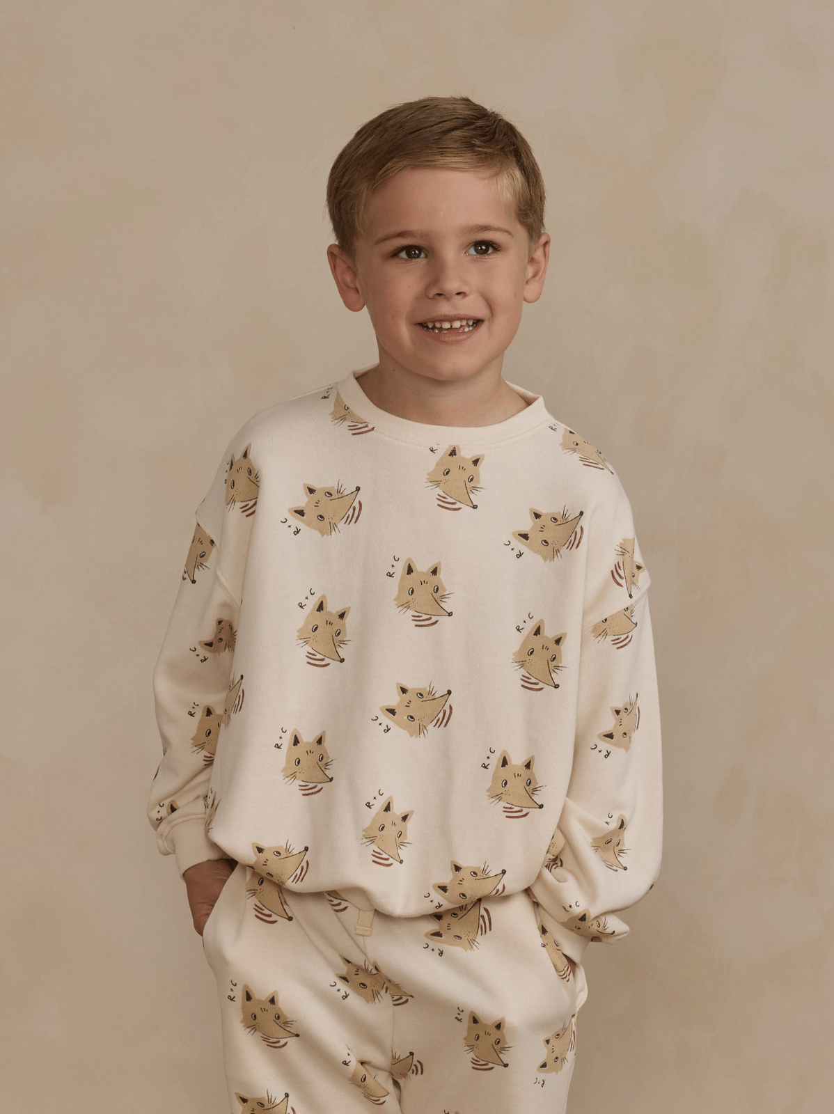 Smiling boy wearing a playful cat-patterned sweatshirt and pants against a neutral background.