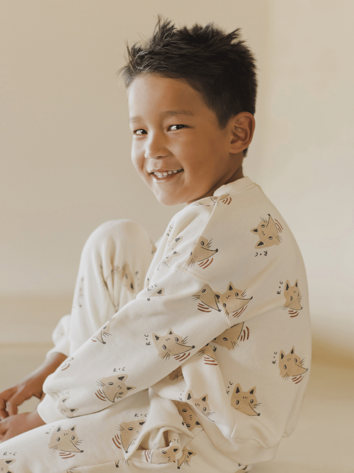 Smiling child in playful cat-patterned pajamas, sitting on the floor against a light background.