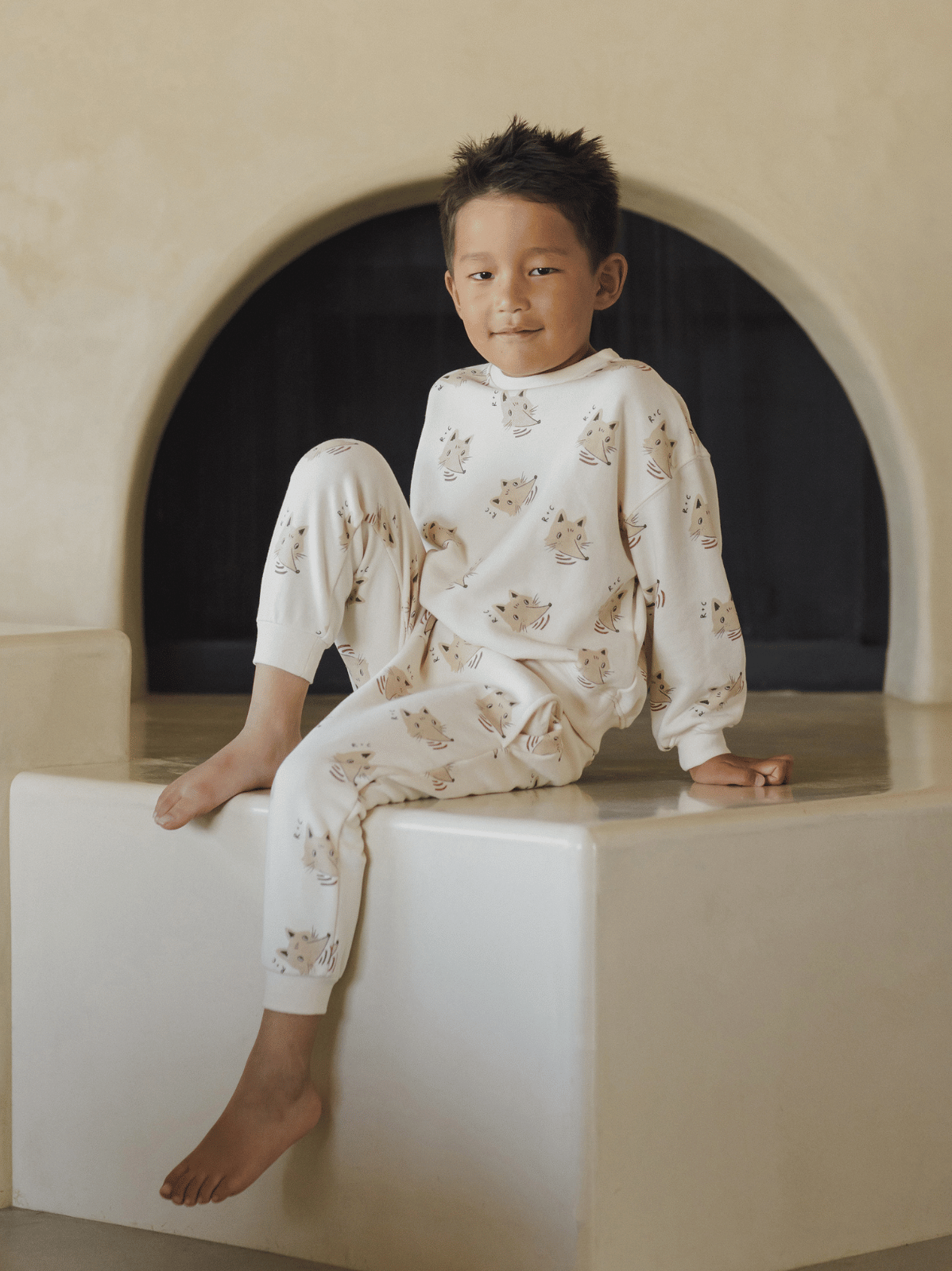 Boy in patterned pajamas sitting on a white pedestal, smiling with an arched wall in the background.