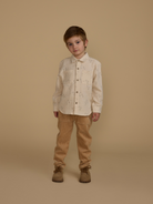 Child standing against a beige background, wearing a light-patterned shirt and tan corduroy pants.