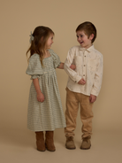 Two smiling children in stylish outfits, a girl in a patterned dress and a boy in a button-up shirt, standing together.