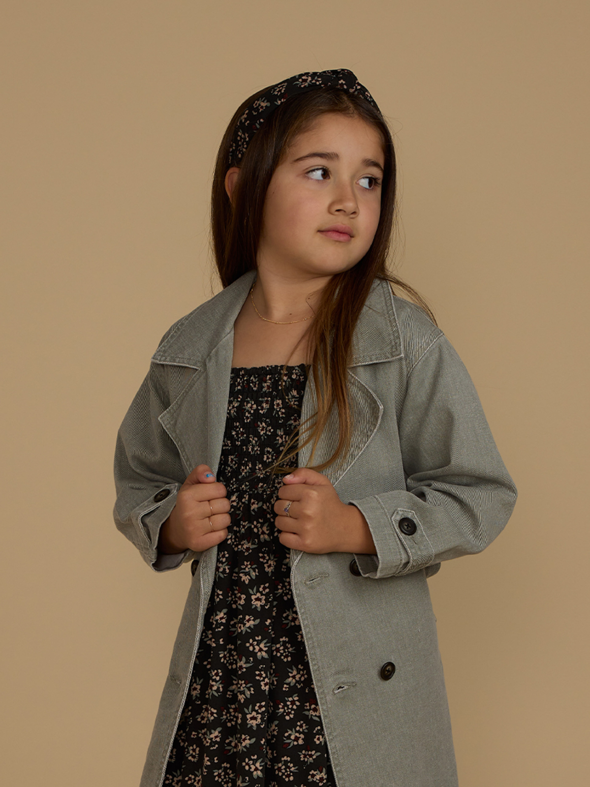 Girl in a trendy gray coat over a floral dress, posing against a beige background.