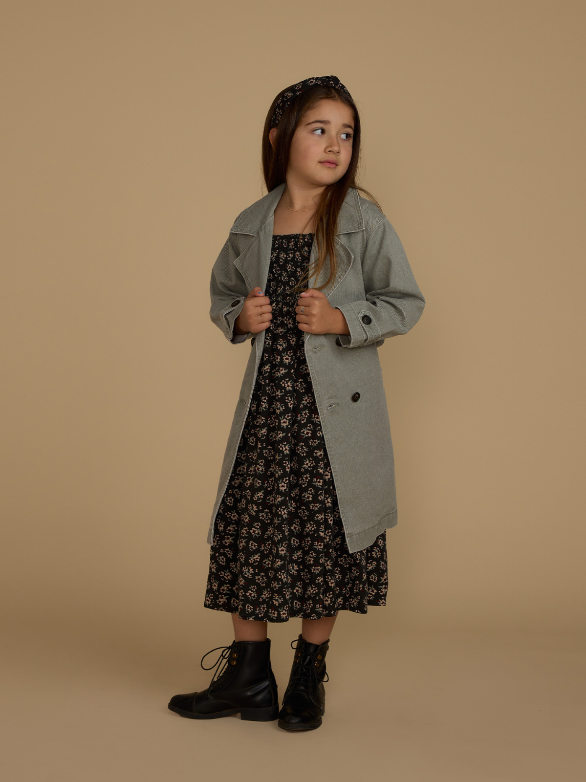 Young girl in a gray coat over a floral dress, standing against a beige background with a thoughtful expression.