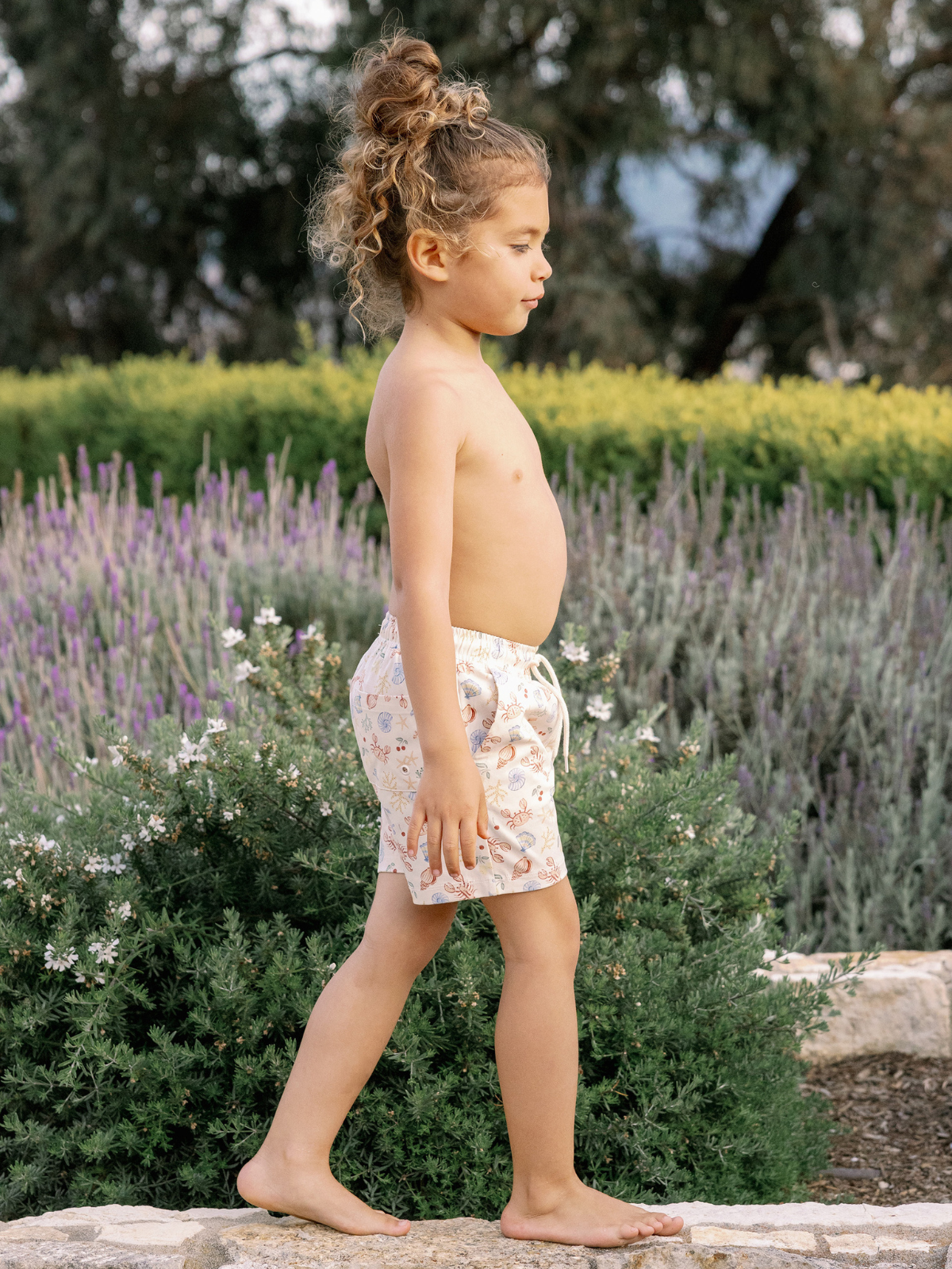Child walking barefoot in a garden, wearing patterned shorts with greenery and lavender in the background.