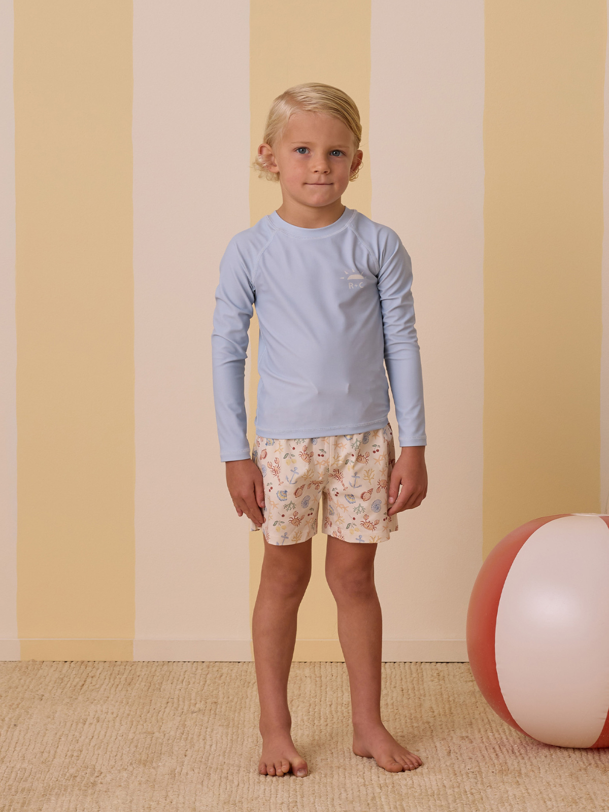 Boy in blue long-sleeve swim top and patterned shorts, standing on a carpet with striped yellow wall and beach ball.