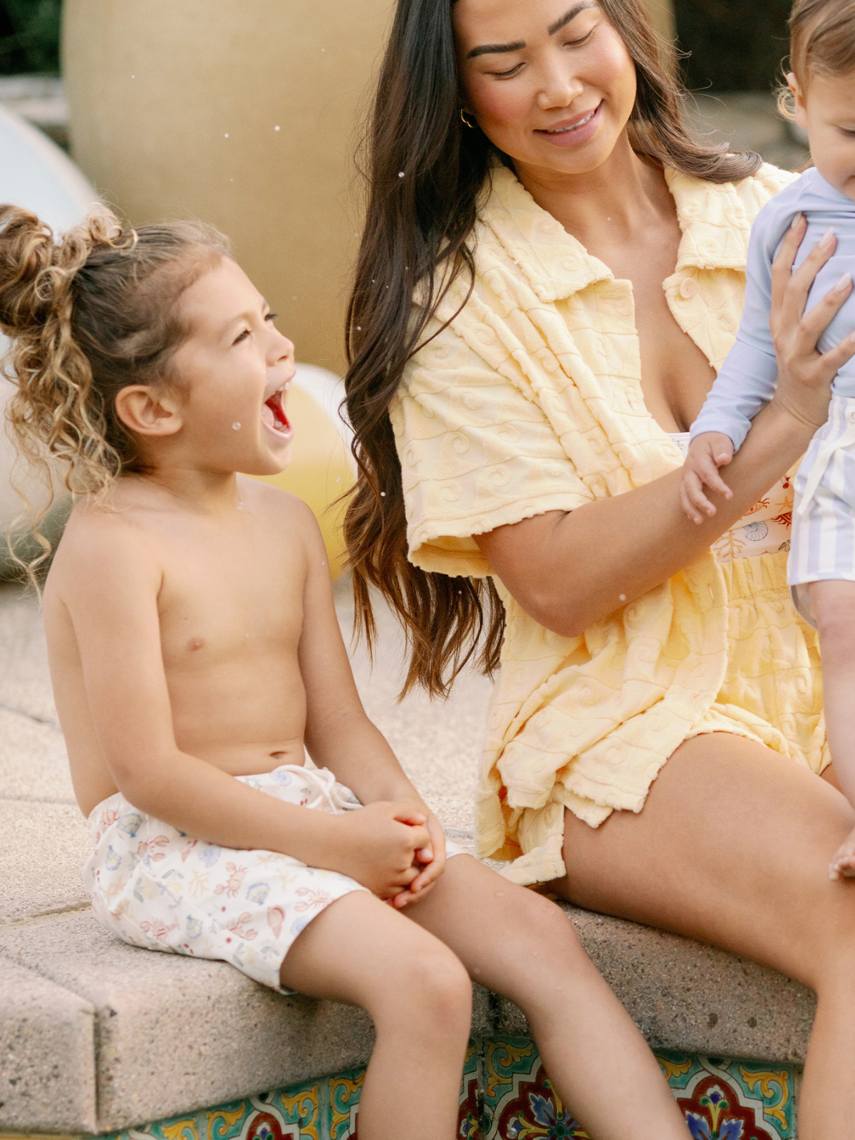 A smiling woman holds a baby while a child laughs beside her, enjoying a sunny day by a pool.
