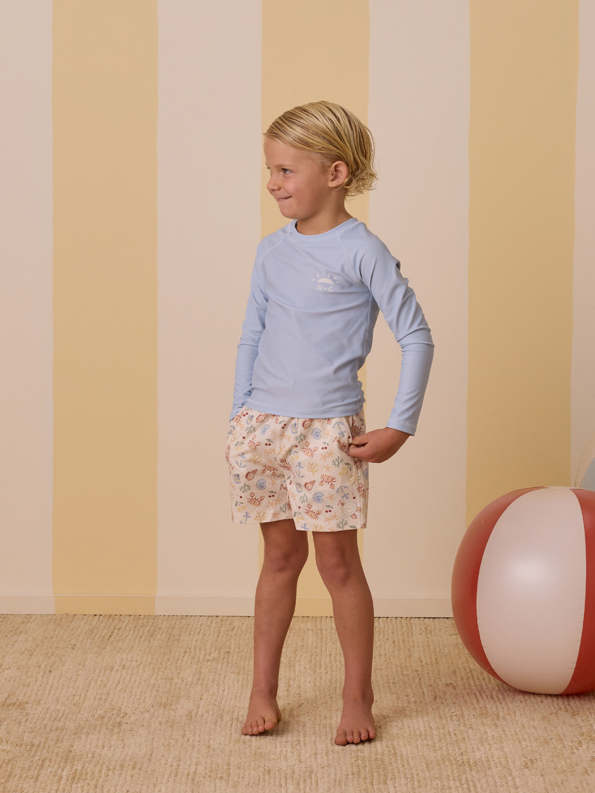 Child standing in swimwear, smiling, against a pastel striped background with a beach ball nearby.
