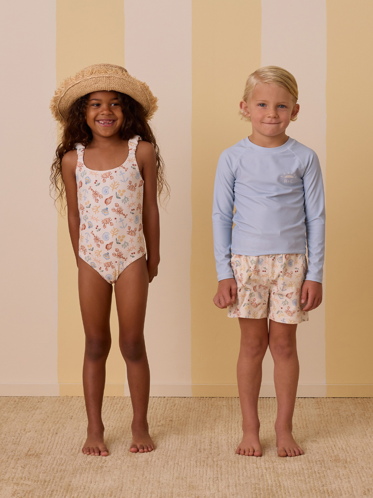 Two children in playful swimwear stand on a light backdrop; one girl in a swimsuit and a boy in a rash guard and shorts.