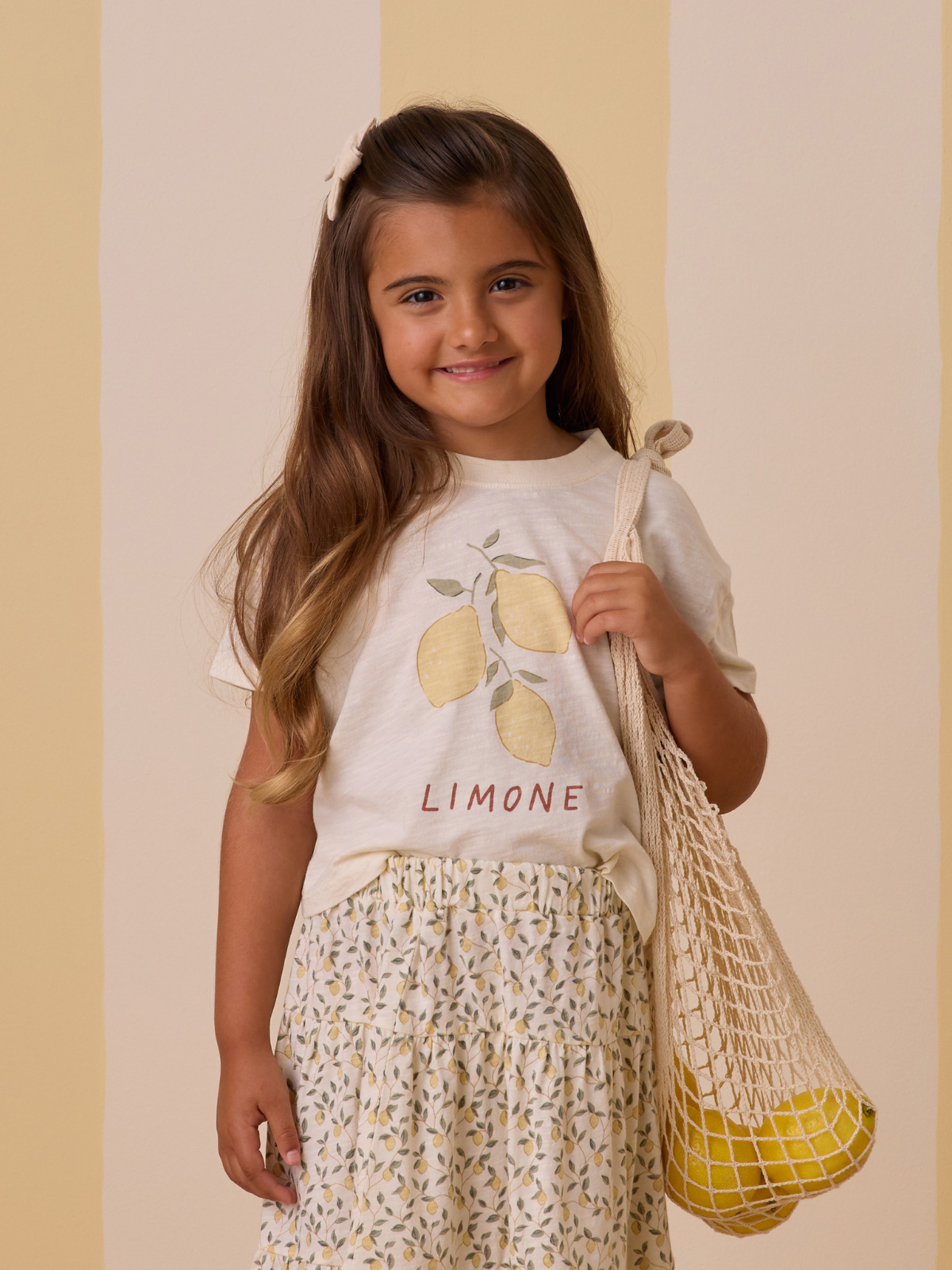 Girl smiling, wearing a lemon graphic tee and floral skirt, holding a net bag with lemons, against a pastel background.