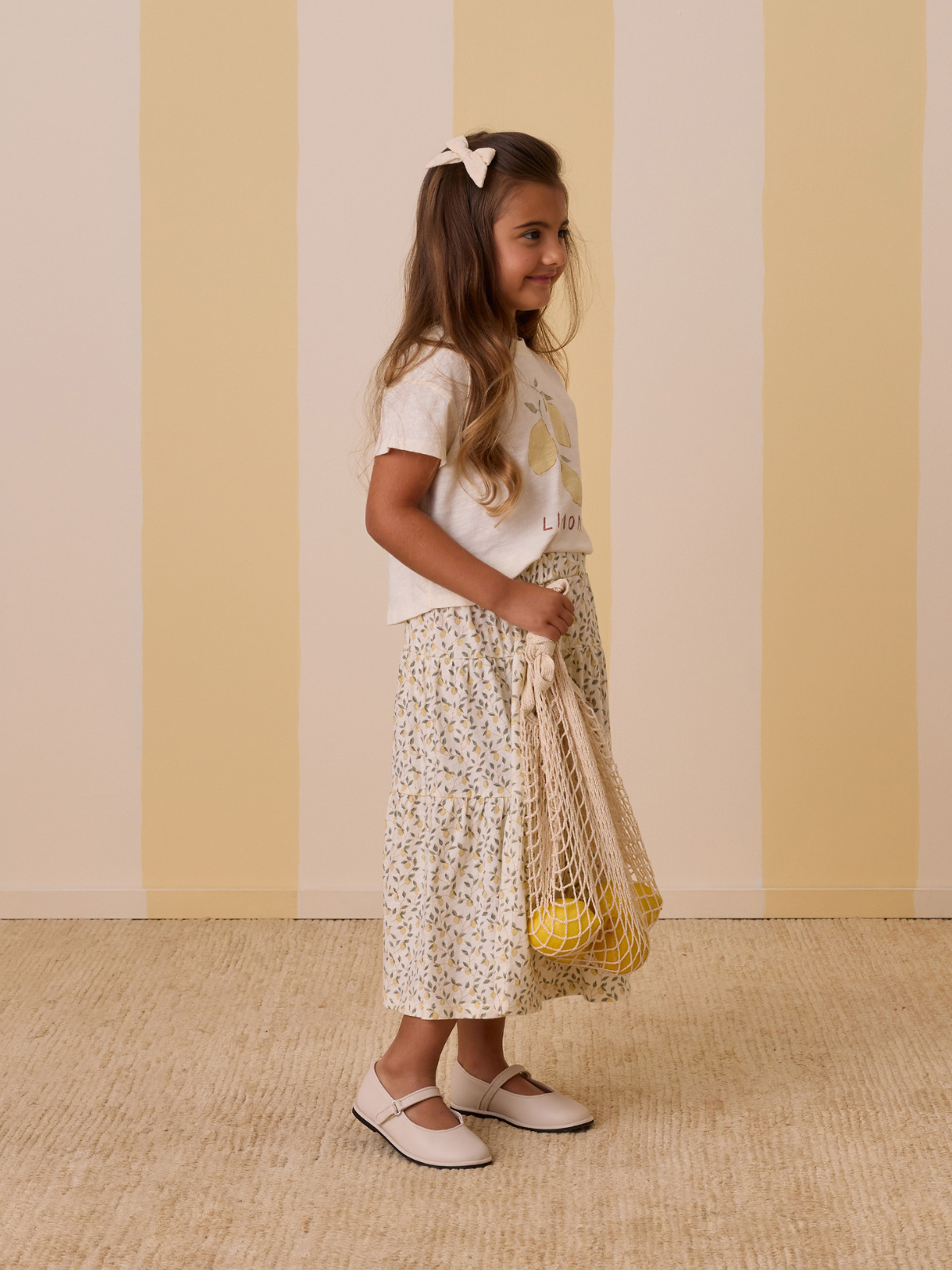 Smiling girl in a lemon-themed shirt, floral skirt, holding a mesh bag with lemons, against a striped background.
