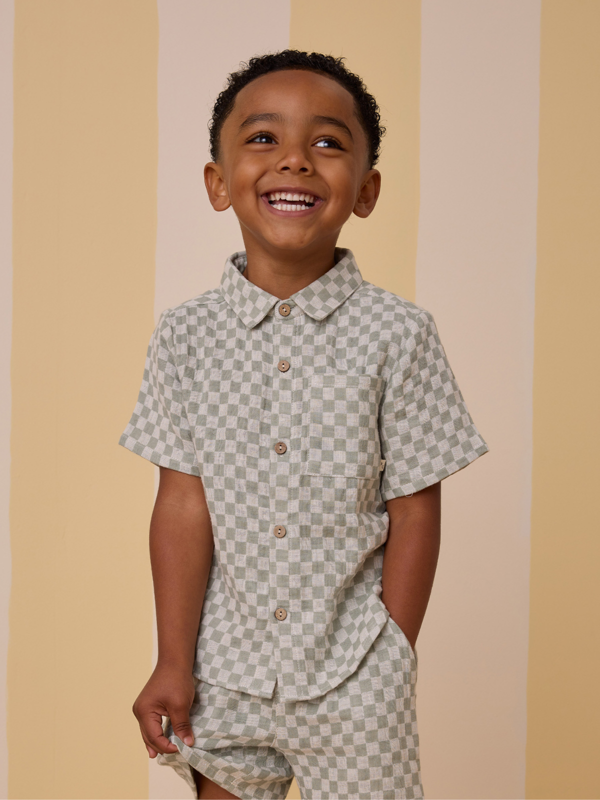Smiling boy in a green and white checkered outfit against a soft yellow striped background.
