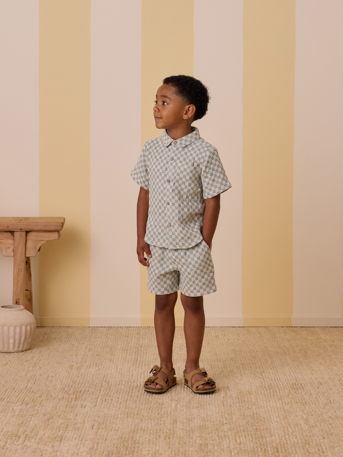 Child wearing a checked short-sleeve shirt and shorts, standing next to a wooden table in a softly striped room.
