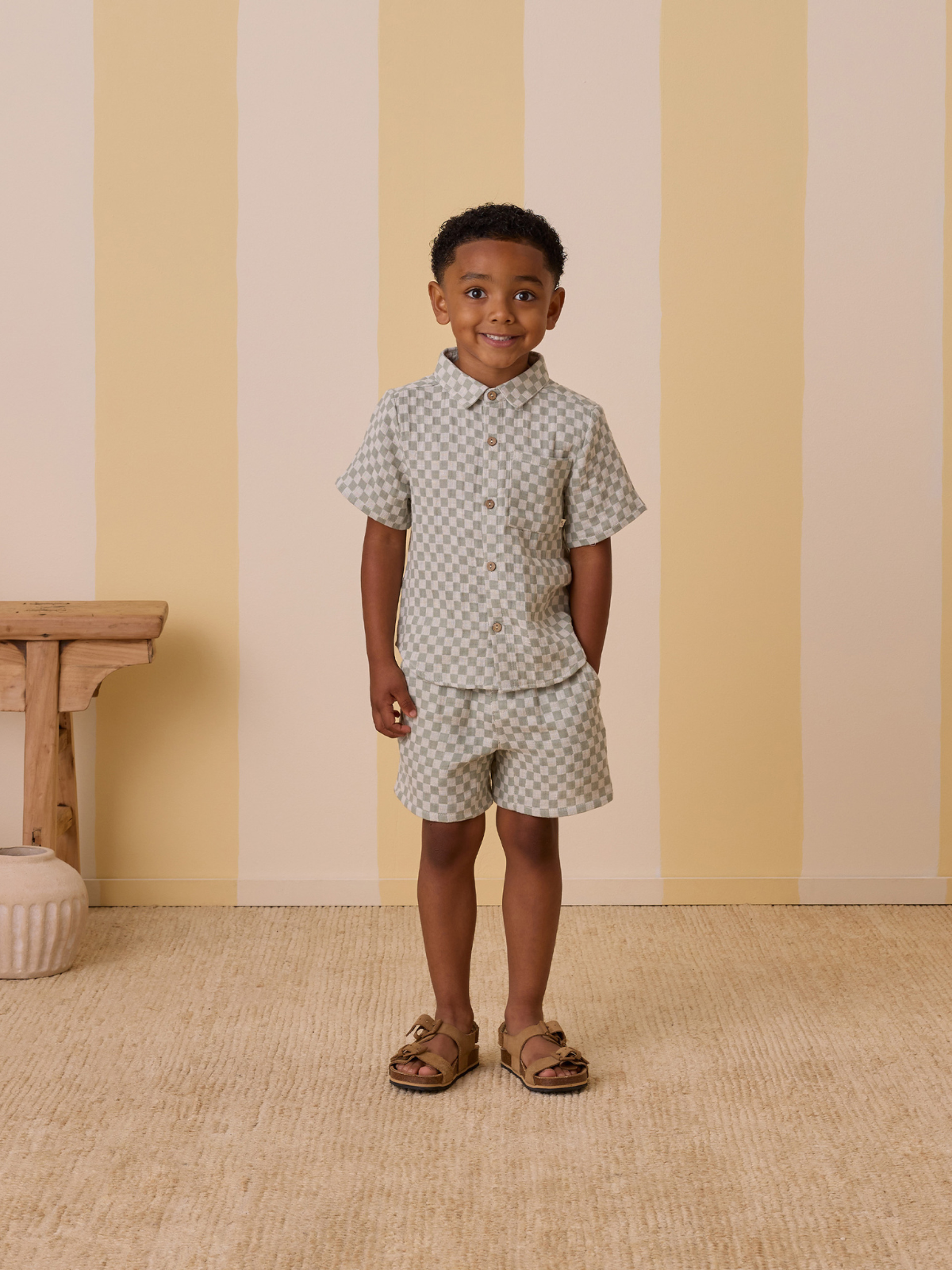 Smiling child in checkered outfit with shorts, standing on a tan rug against a striped wall.