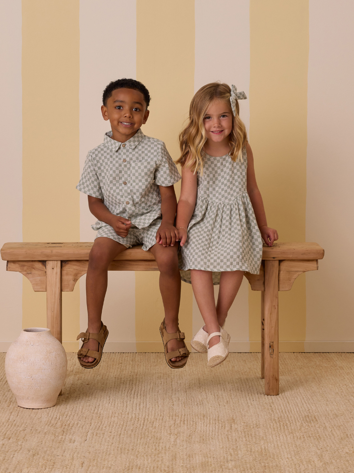 Two smiling children sit on a wooden bench, wearing matching checker-patterned outfits against a striped backdrop.
