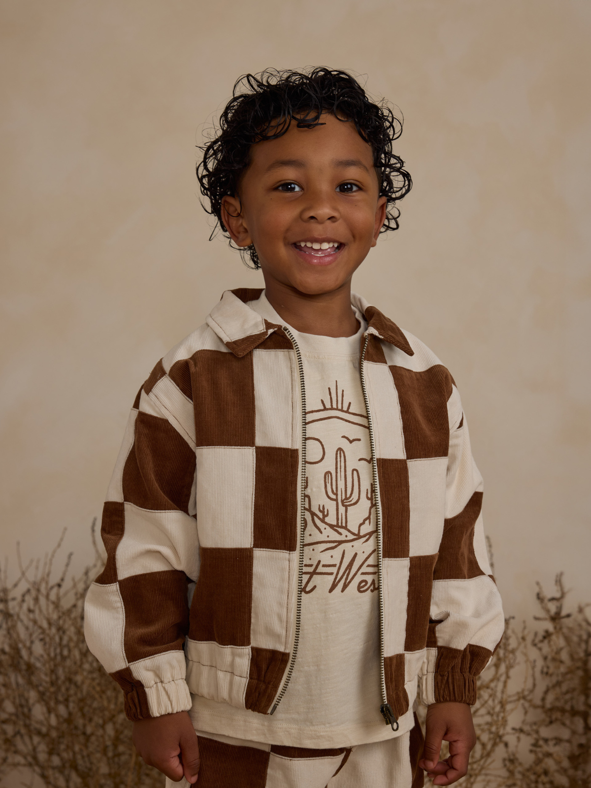 Smiling child in a checkered brown and cream jacket against a textured background, promoting a playful vibe.