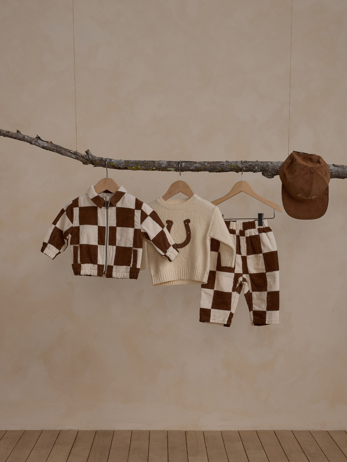 Children's winter clothing hanging on a branch: brown and cream checkered jacket, sweater, pants, and cap.