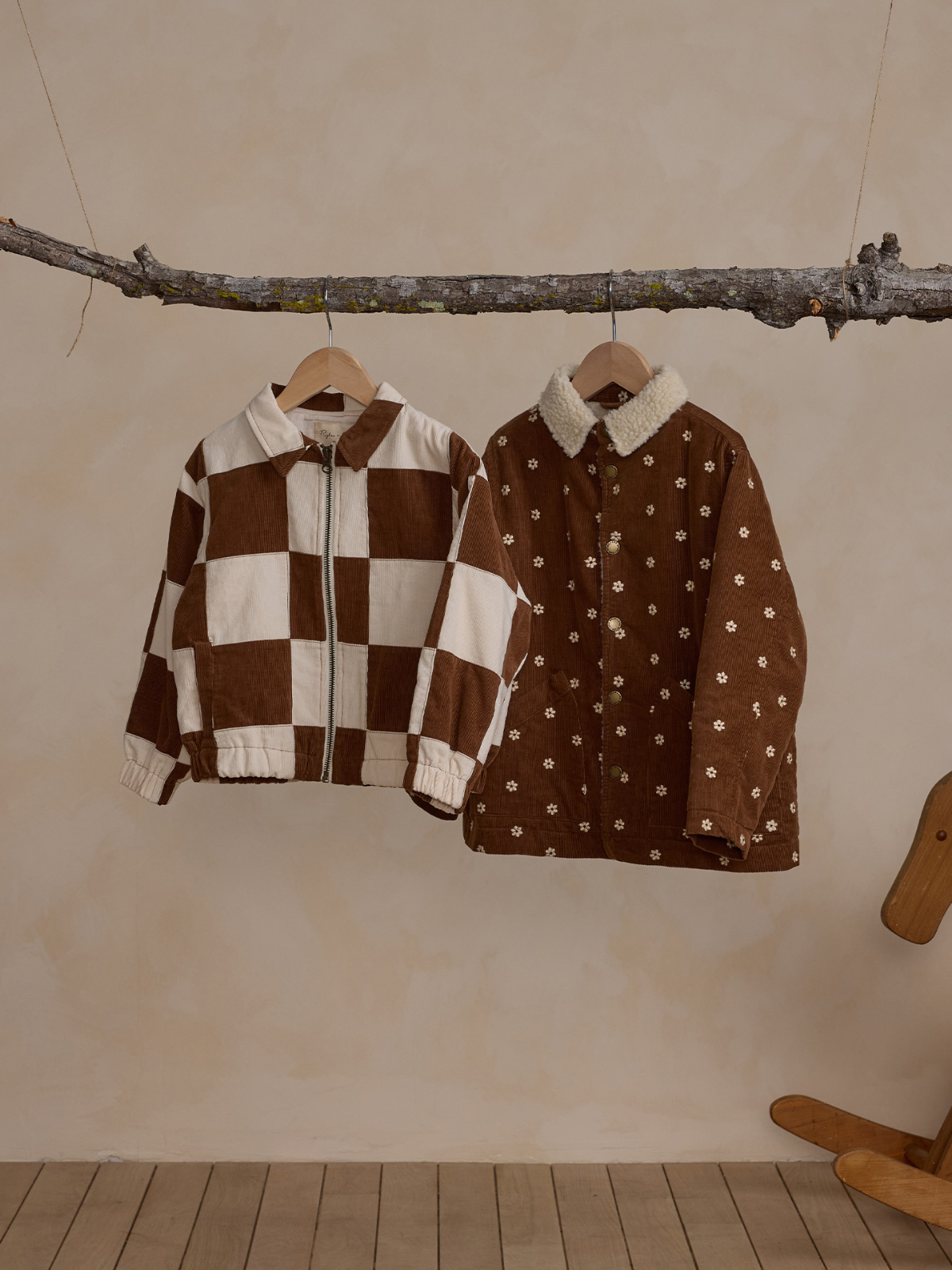 Two stylish jackets hang from a branch: a brown and white checkered design and a brown floral embroidered coat.