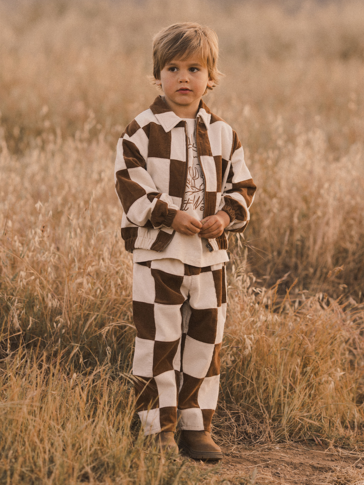 Child in checkered brown and white outfit stands in a field during sunset, surrounded by tall grass.