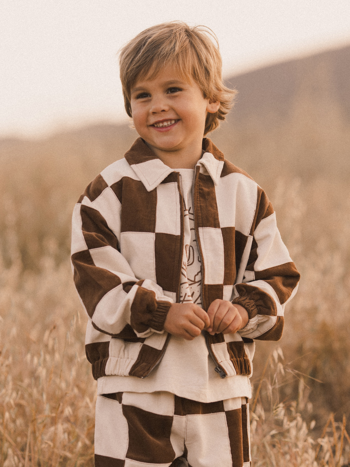 Smiling child in a brown and white checkered jacket and matching pants, standing in a field during sunset.