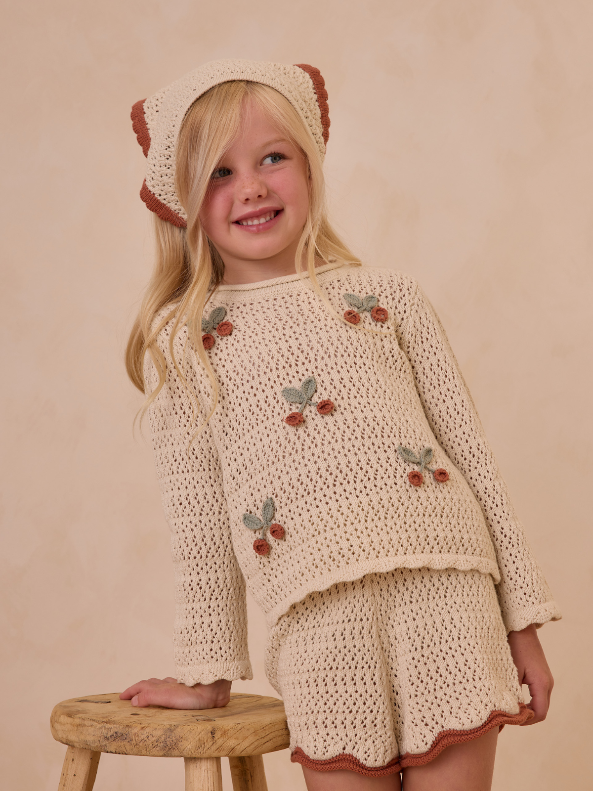 Smiling girl in a knitted outfit with flowers, standing beside a wooden stool against a light background.