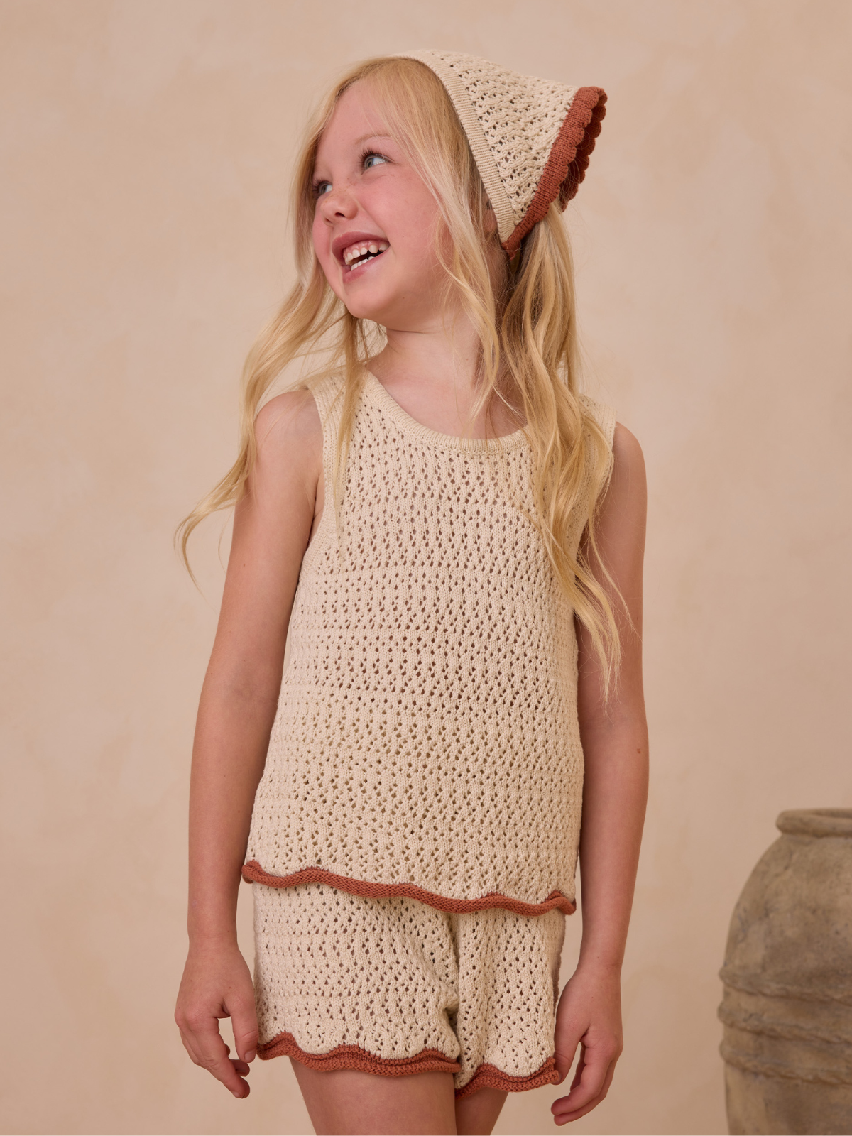 Smiling girl in a cream knit outfit with a brown-trimmed hat, standing against a soft, neutral background.