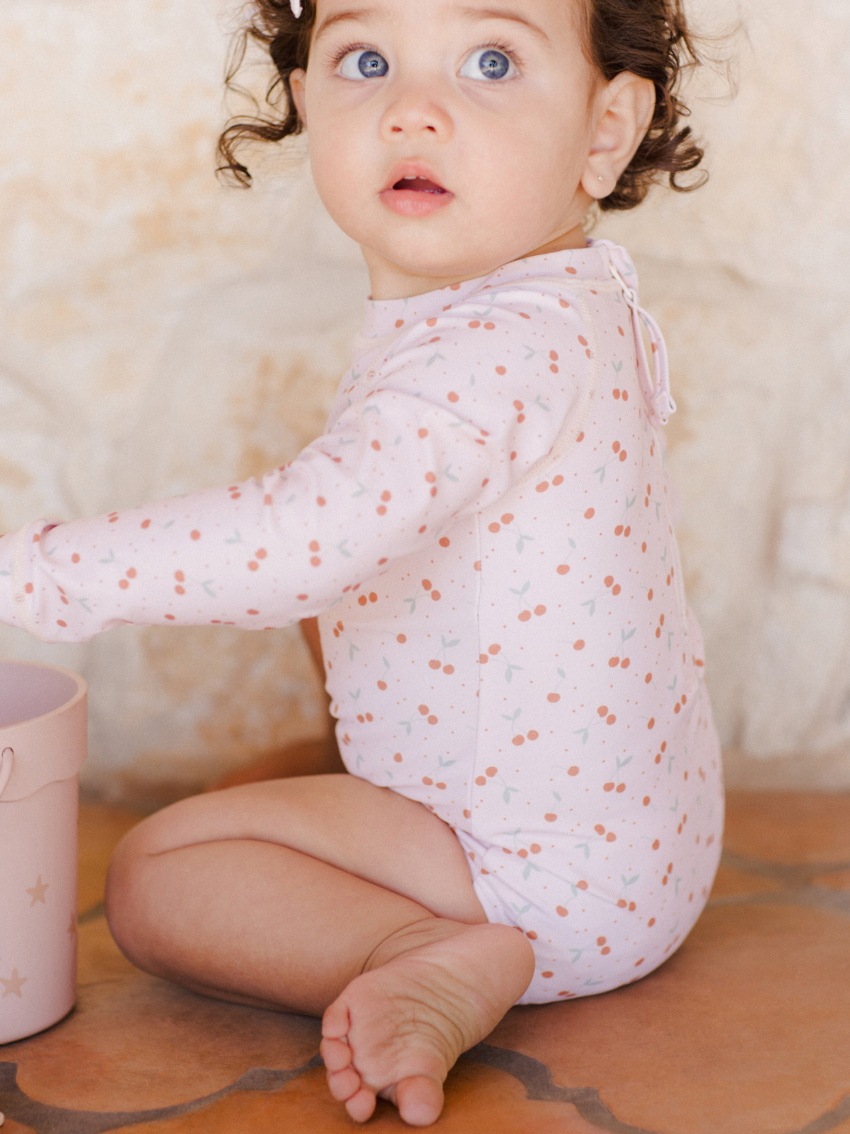 Baby with curly hair in a pink patterned onesie, looking surprised, sits on a tiled floor near a pink bucket.