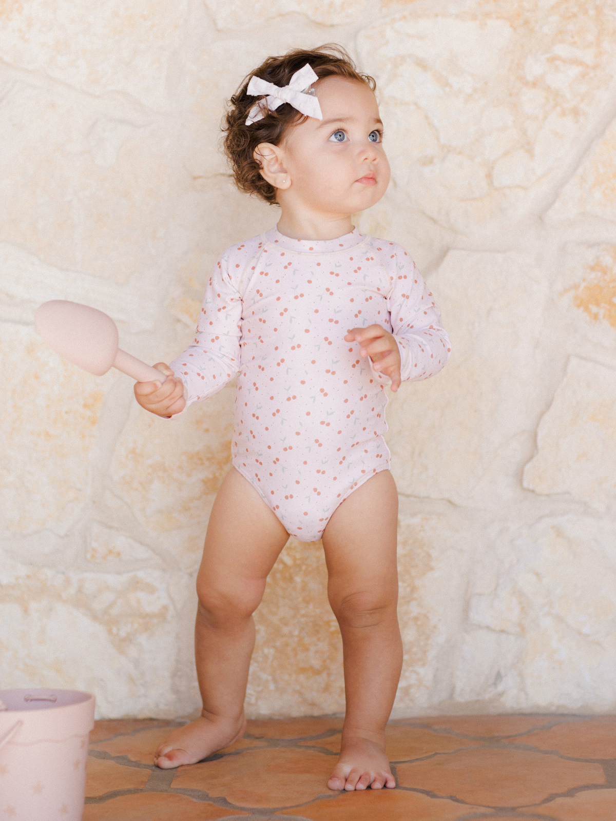 Toddler in a pink patterned swimsuit and bow, holding a toy against a stone wall background.