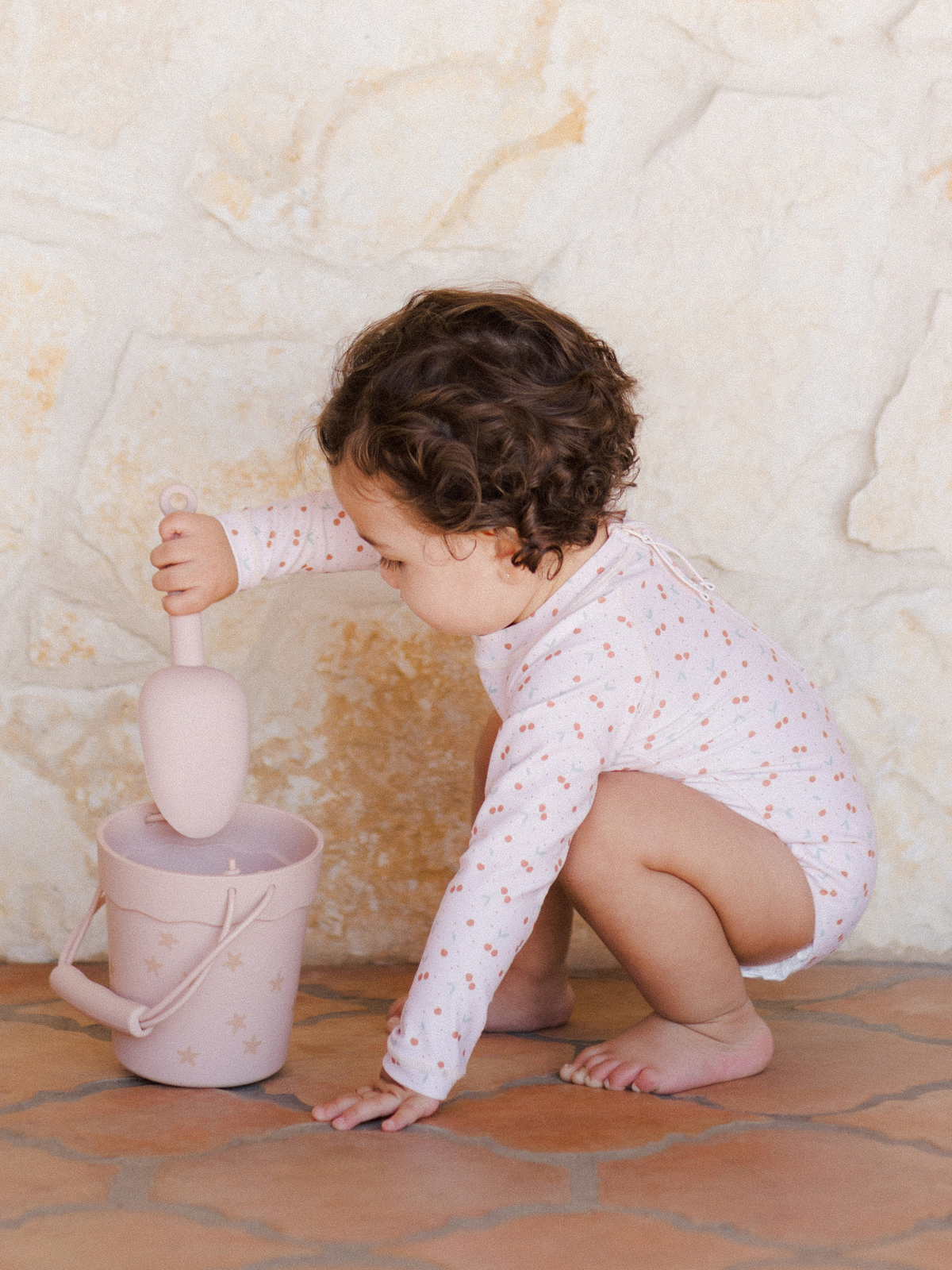 Child in a polka dot onesie playing with a pink bucket and shovel on a textured floor.