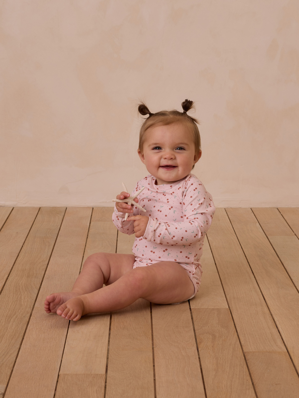 Smiling baby sitting on wooden floor, wearing a pink long-sleeve onesie with colorful dots.
