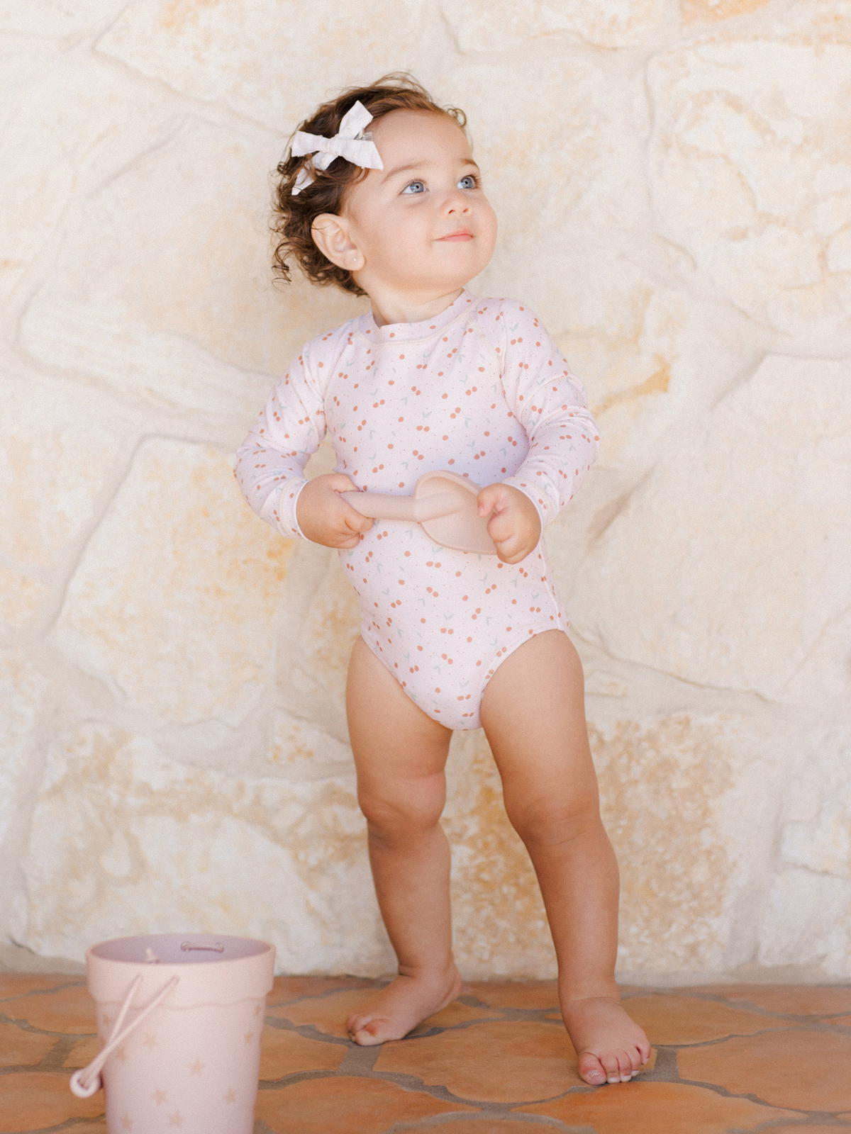 Toddler in a star-patterned bodysuit holds a pink shovel, with a matching bucket, standing against a stone wall.