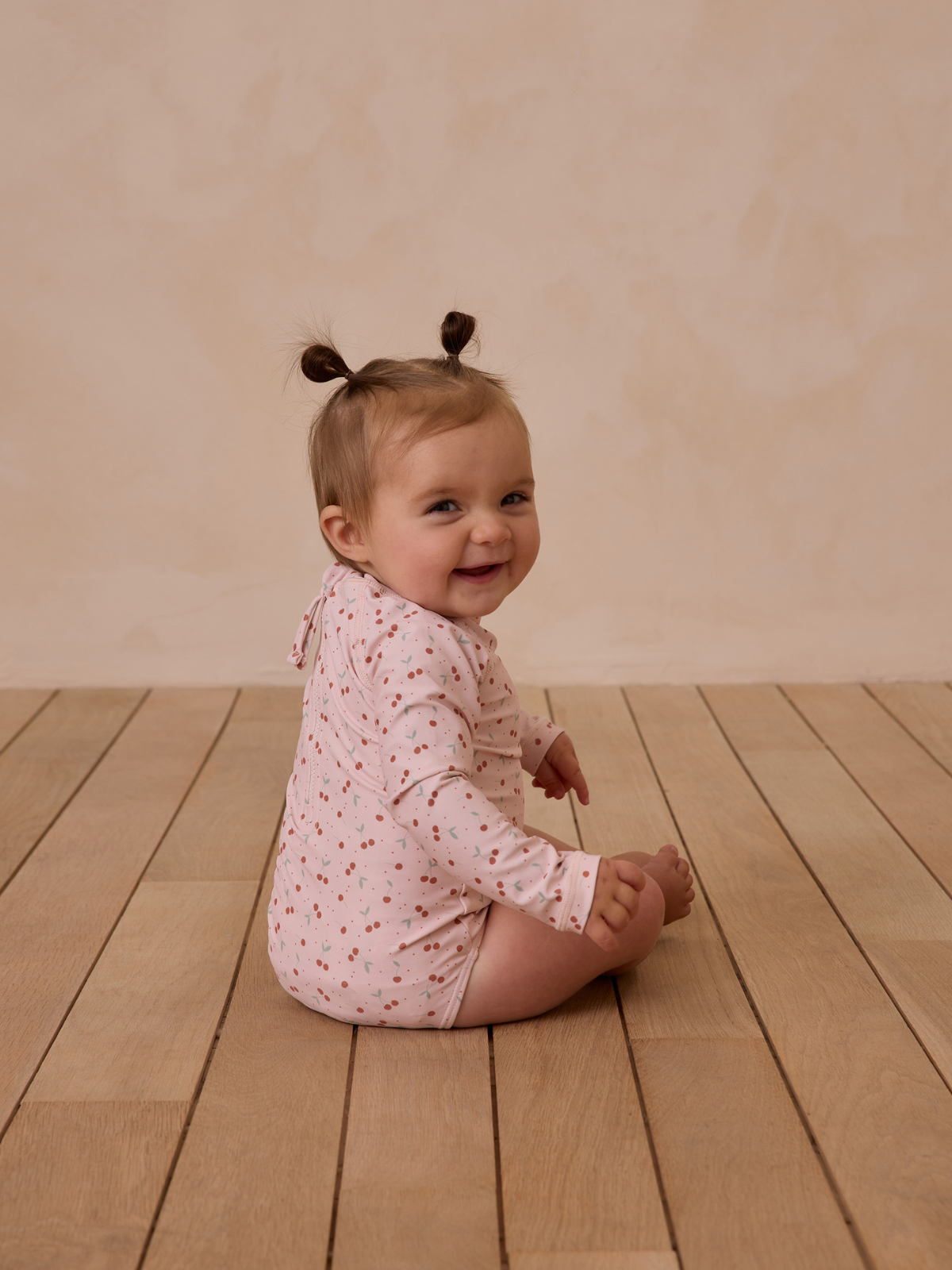 Smiling baby with two ponytails, sitting on a wooden floor, wearing a pink patterned outfit.