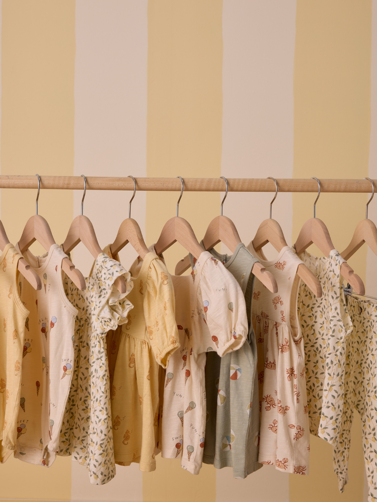 A display of colorful children's dresses hanging on wooden hangers against a striped yellow wall.