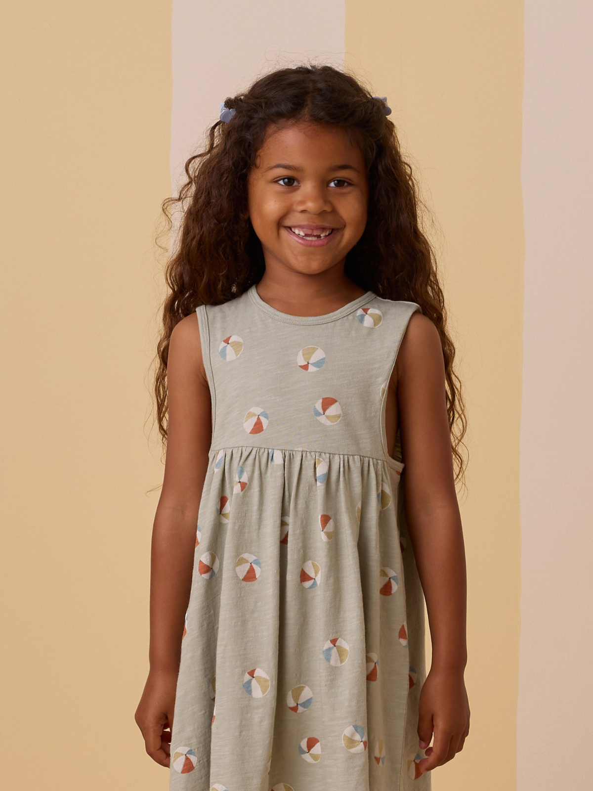 Smiling girl with long curly hair wearing a light dress with colorful patterns, standing against a soft pastel background.