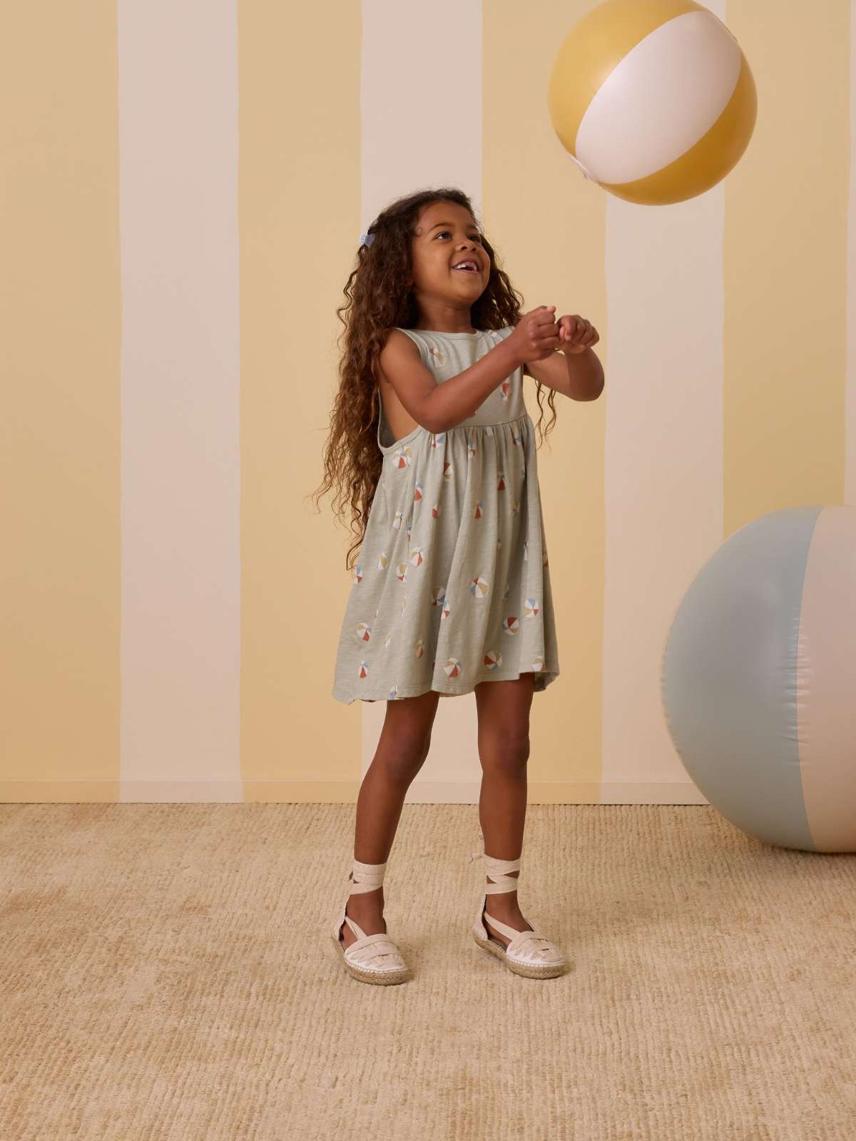 Child in a playful dress smiles while tossing a beach ball in a bright, striped room with pastel colors.