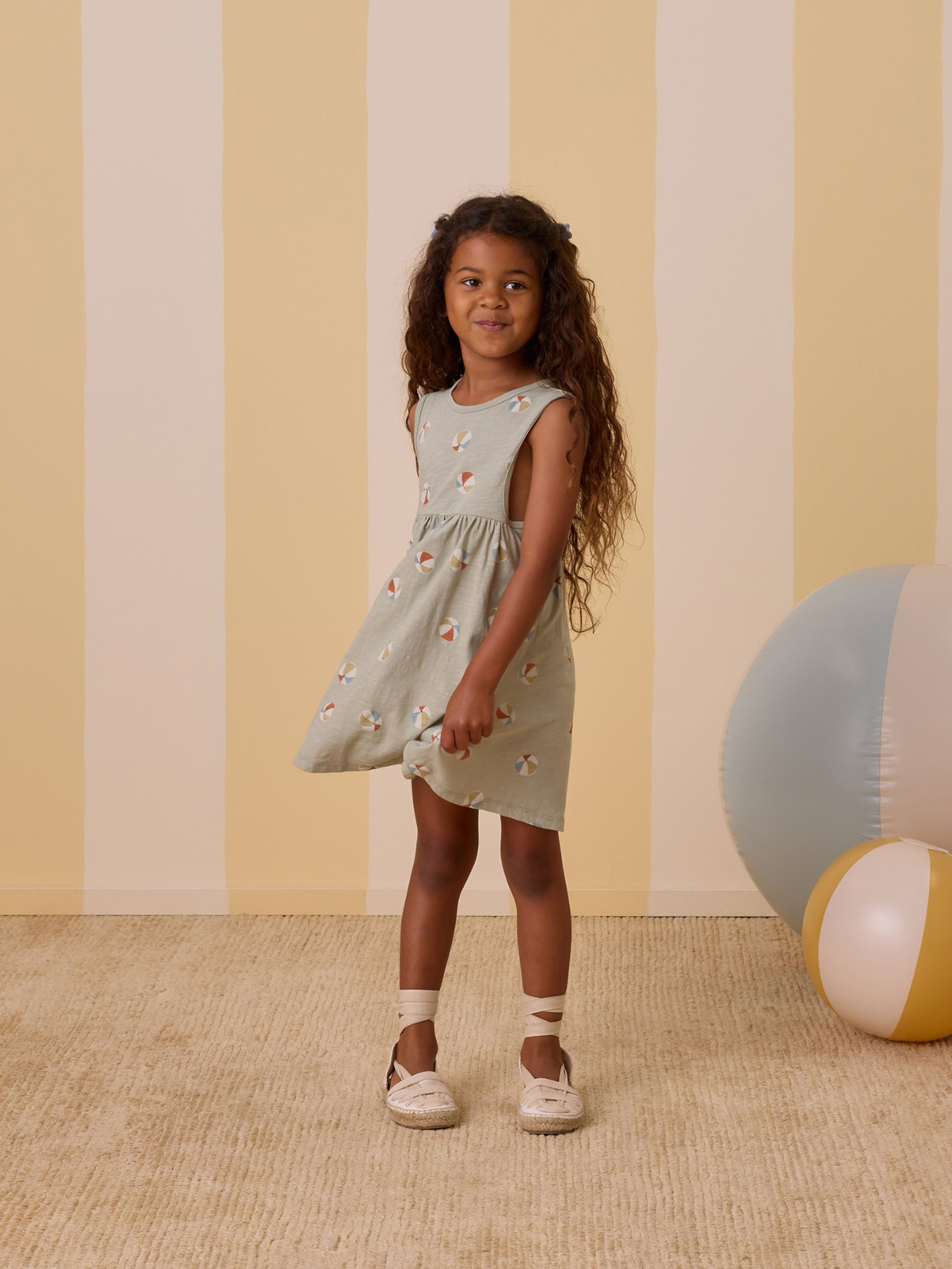 Smiling girl in a patterned dress stands on a carpet, surrounded by colorful beach balls and striped walls.