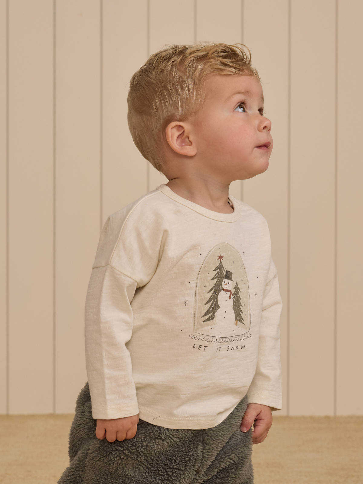 Toddler in a light sweater with a snowman design, looking up against a soft, neutral background.