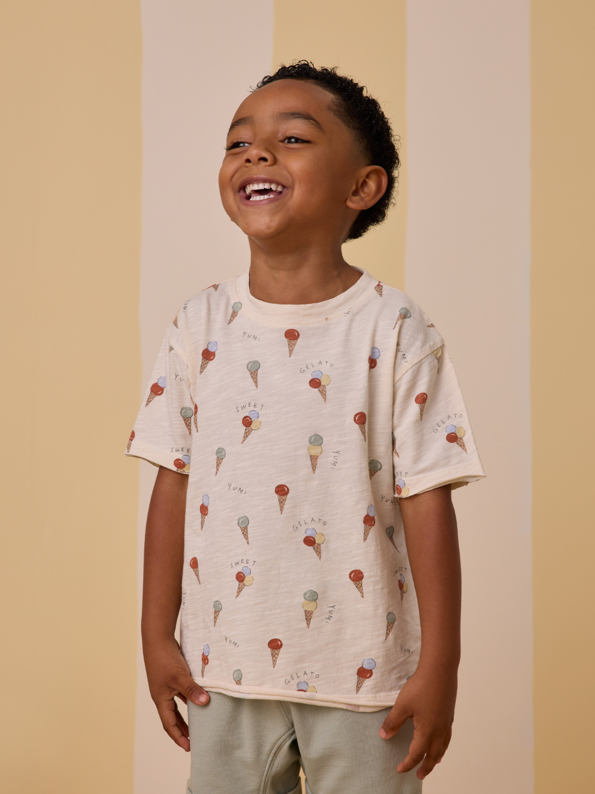 Smiling child wearing a cream tee with colorful ice cream cone patterns, against a soft striped background.