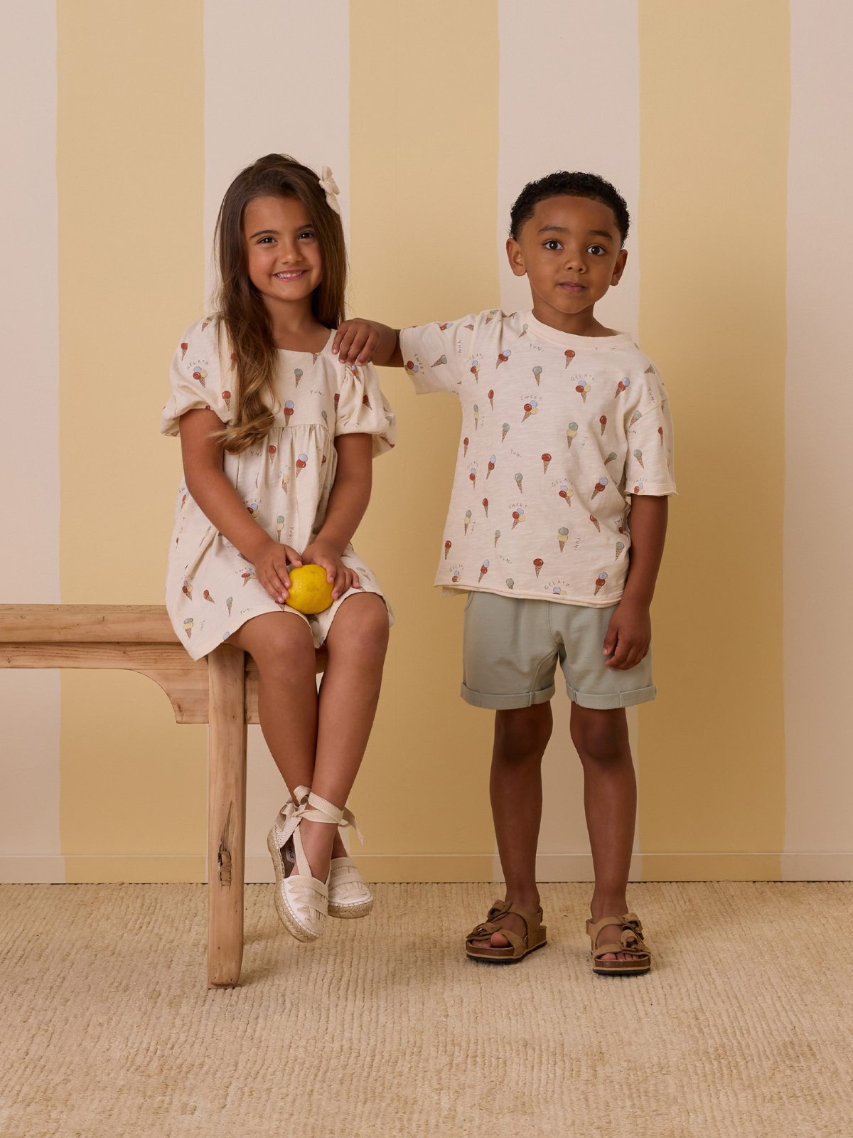 Two children pose in summer outfits, one holding a lemon, on a wooden bench against a striped background.
