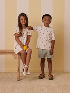 Two children pose in summer outfits, one holding a lemon, on a wooden bench against a striped background.