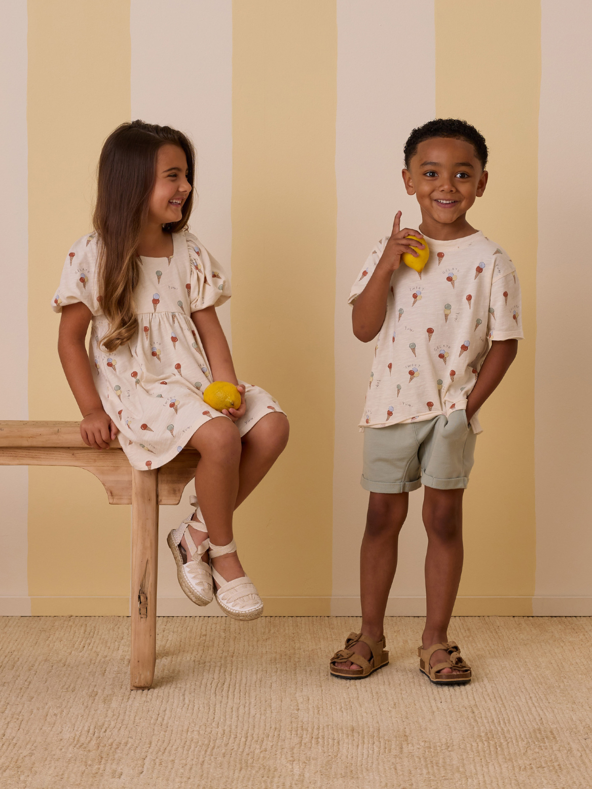 Two children smile while posing for a photo, dressed in playful summer outfits against a striped backdrop.