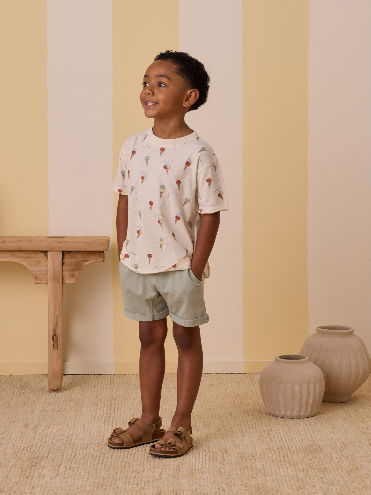 Smiling boy in ice cream print shirt and shorts, standing against a pastel striped wall with decorative vases.