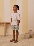 Smiling boy in ice cream print shirt and shorts, standing against a pastel striped wall with decorative vases.