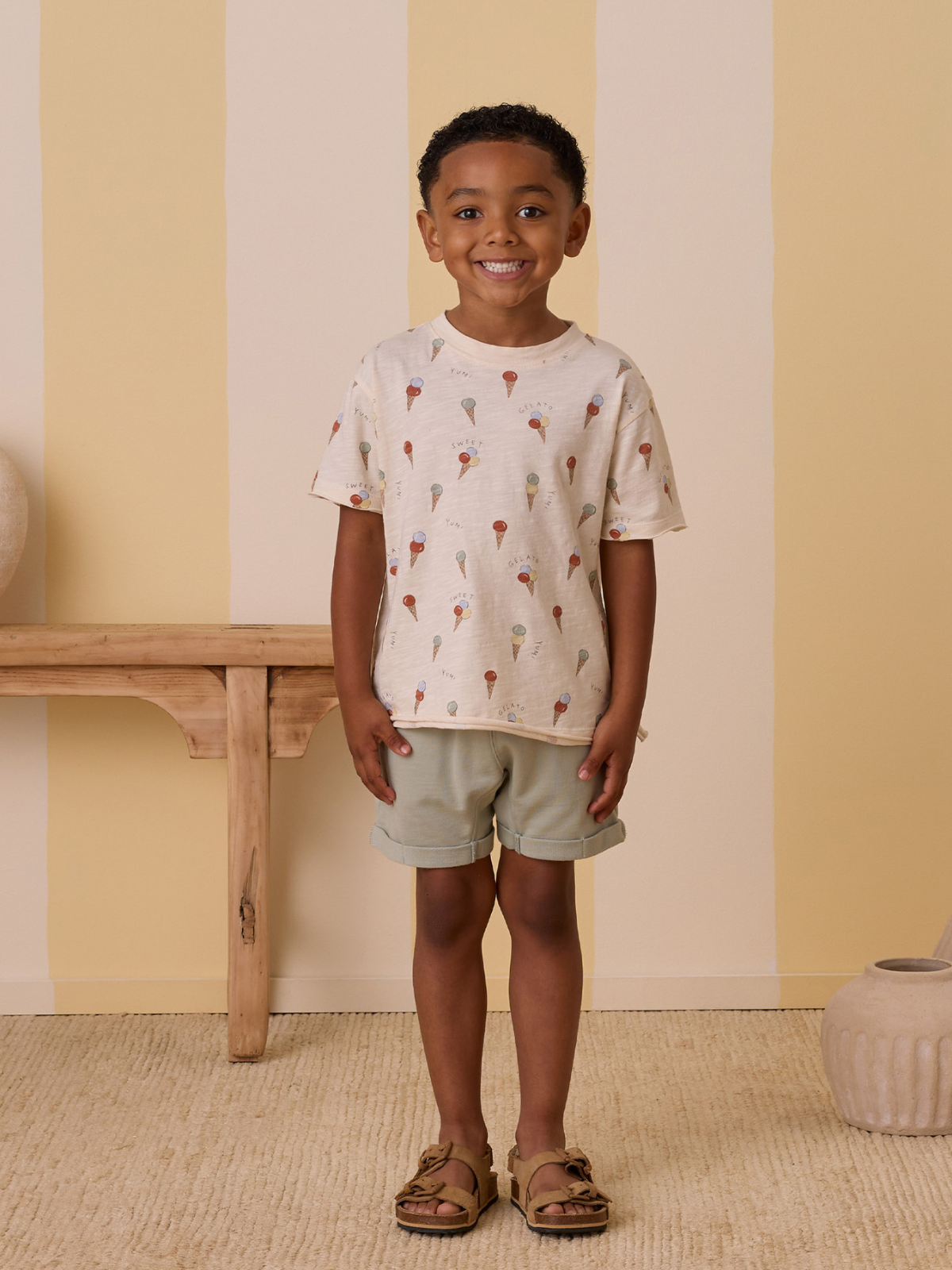 Smiling child with curly hair in a playful ice cream print shirt, light shorts, and sandals, standing indoors.