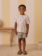 Smiling child with curly hair in a playful ice cream print shirt, light shorts, and sandals, standing indoors.