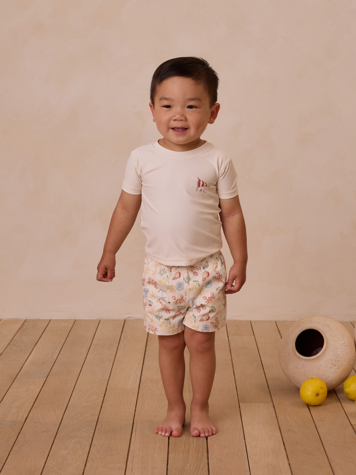 Smiling young boy in a light shirt and patterned shorts, standing on wooden floor with a neutral backdrop.