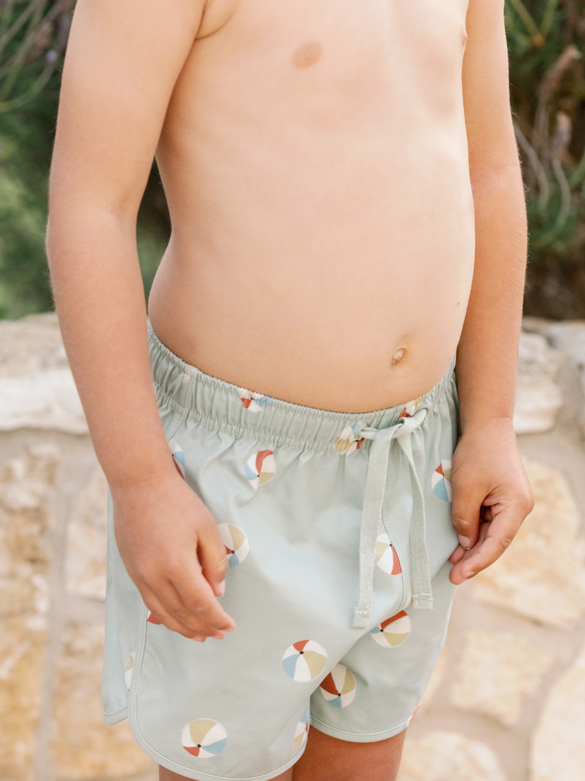 Child wearing light green swim shorts with a beach ball pattern, standing by a stone wall.